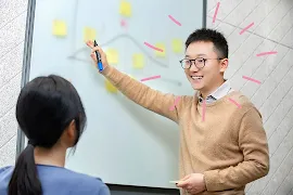 Boy in focus, wearing a brown sweater and writing on whiteboard, looking at another person.